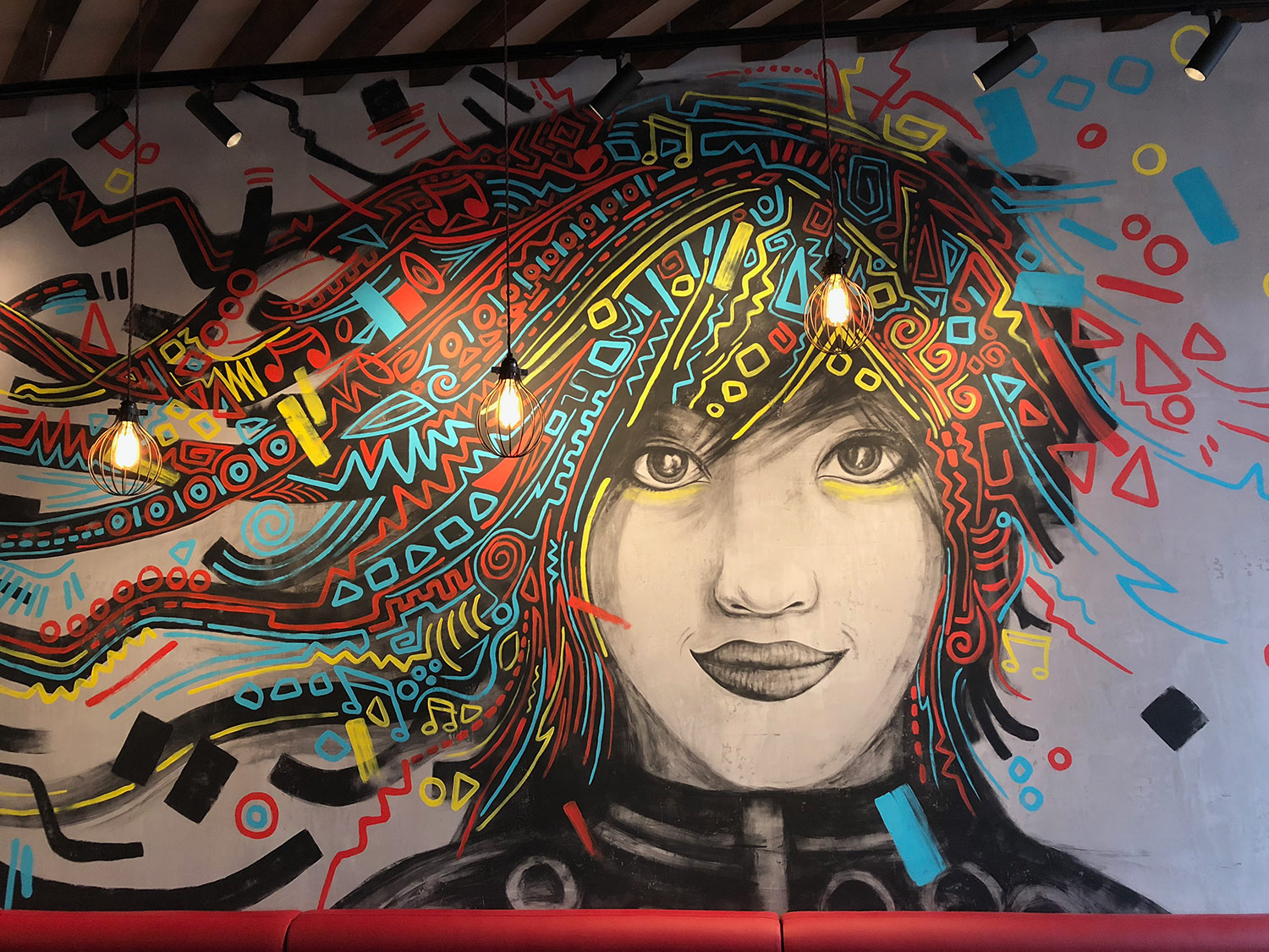 A mural of a portrait of a smiling girl with black, red, blue and yellow doodles for hair on a grey wall, in MOD Pizza, Leicester by SAINT Design.