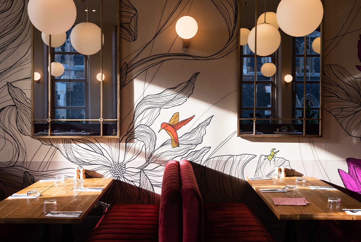 Interior design of a restaurant, showing seating booths and a detailed mural of flowers, plants and birds painted direct to the walls. Mural by SAINT Design for mallow, a vegan restaurant in London.