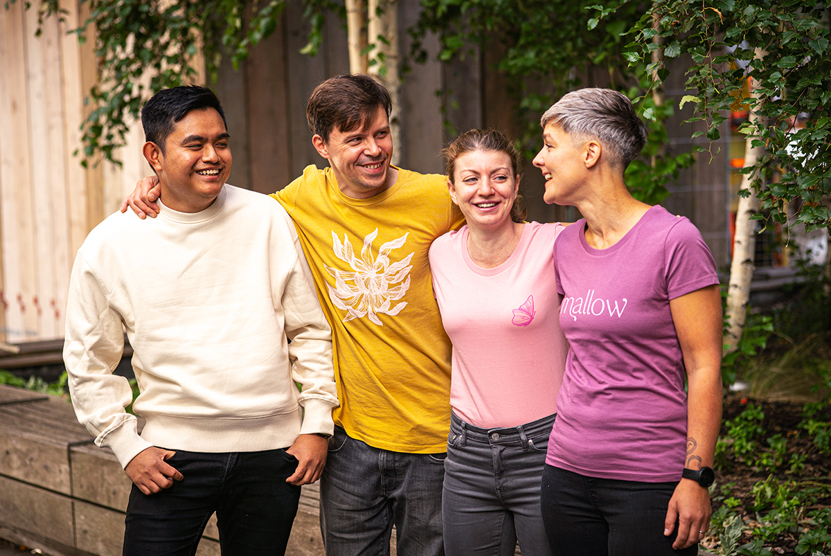 Four people in uniforms. Various sweatshirts and t-shirts, one cream and plain, one yellow with a pink illustration of a flower, one is pink with a darker pink butterfly illustration and one dark with light brand wordmark. By SAINT Design for mallow, a vegan restaurant in London.