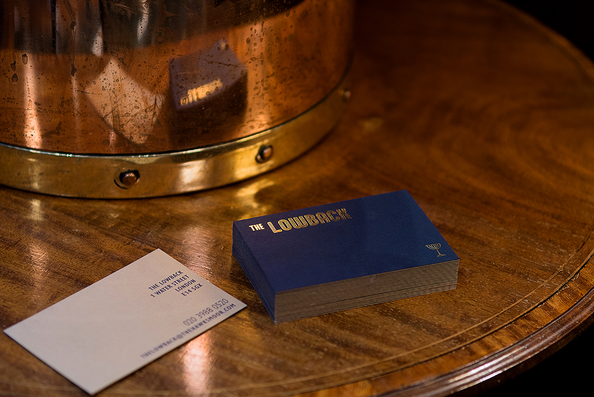 Stack of business cards with The Lowback logo in gold foil as a design detail photographed on a dark wood table top, by SAINT Design for Hawksmoor.