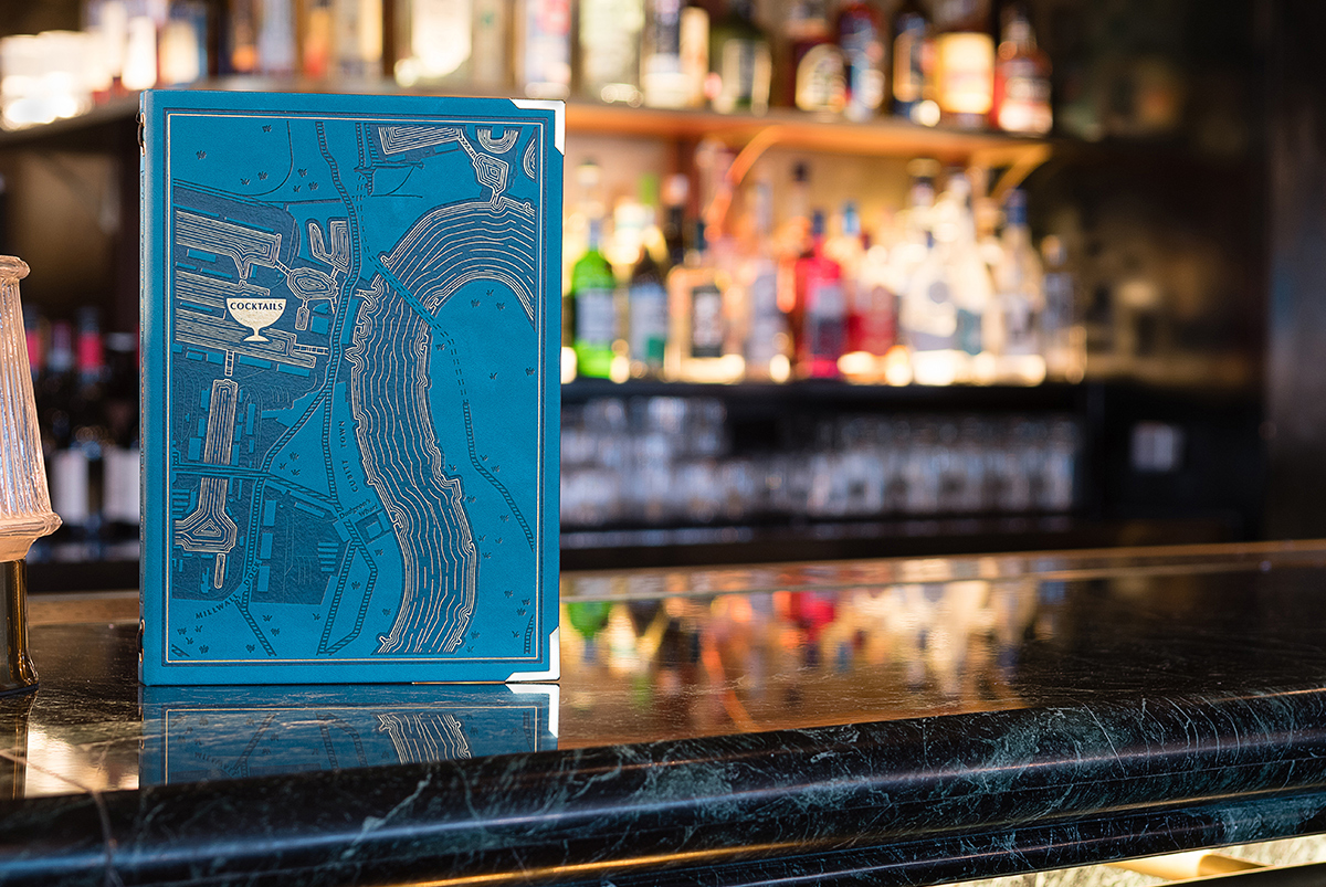The front of a blue leather-like menu cover with a map of the Isle of Dogs as a design detail photographed on the bar top, by SAINT Design for Hawksmoor.